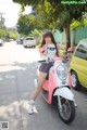 A woman sitting on a pink scooter on a city street.