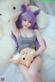 A woman with purple hair is sitting on a bed with a teddy bear.