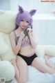 A woman with purple hair sitting on a bed next to a teddy bear.