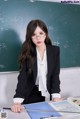 A woman in a black suit and glasses sitting at a desk in front of a blackboard.